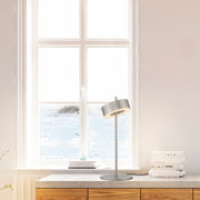 Moderne Tischleuchte Q-ROTATE in silberner Farbe, seitliche Perspektive, auf einem Tisch vor einem Fenster platziert.