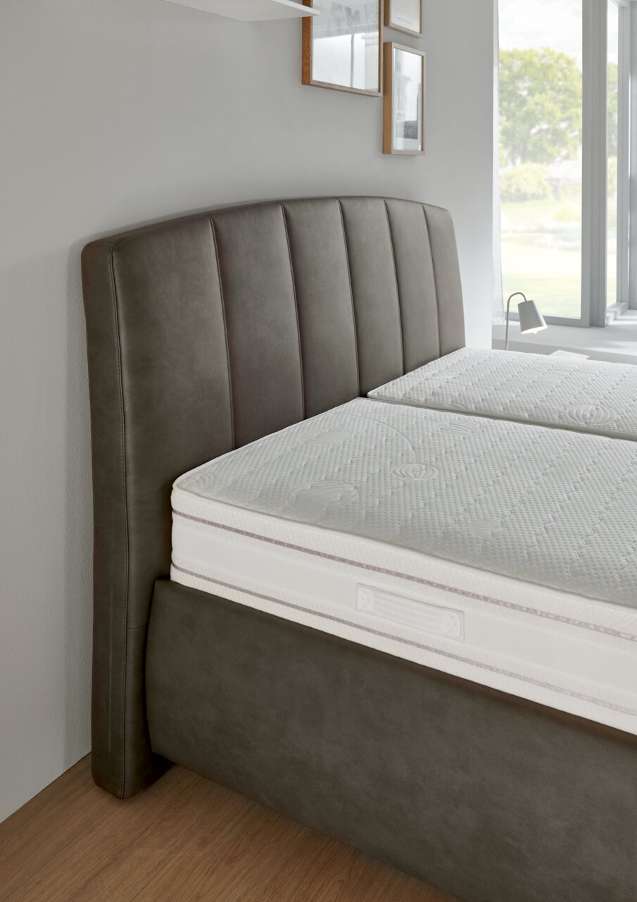 Seitliche Perspektive eines Polsterbetts mit gepolstertem Kopfteil in dunklem Stoff und einer weißen Matratze.