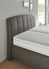 Seitliche Perspektive eines Polsterbetts mit gepolstertem Kopfteil in dunklem Stoff und einer weißen Matratze.