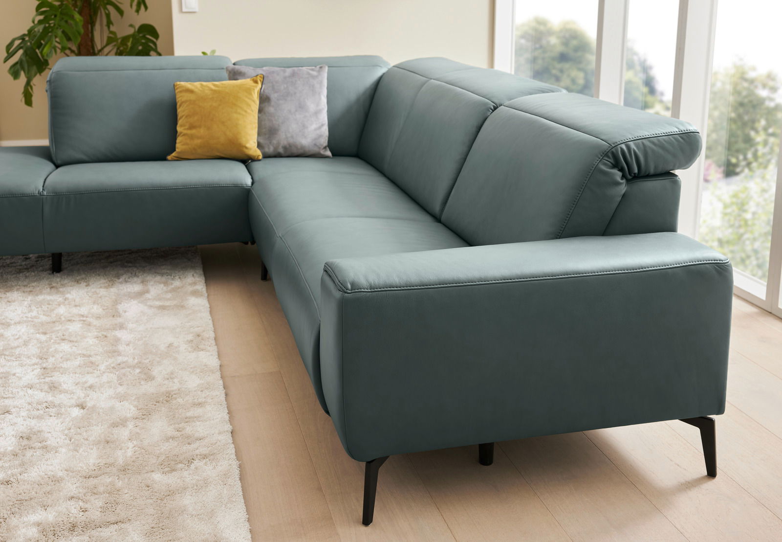 Eckgarnitur in modernem Design mit blauen Polstern, gelbem und grauem Kissen, auf hellem Holzboden und vor großen Fenstern, Perspektive von schräg vorne.