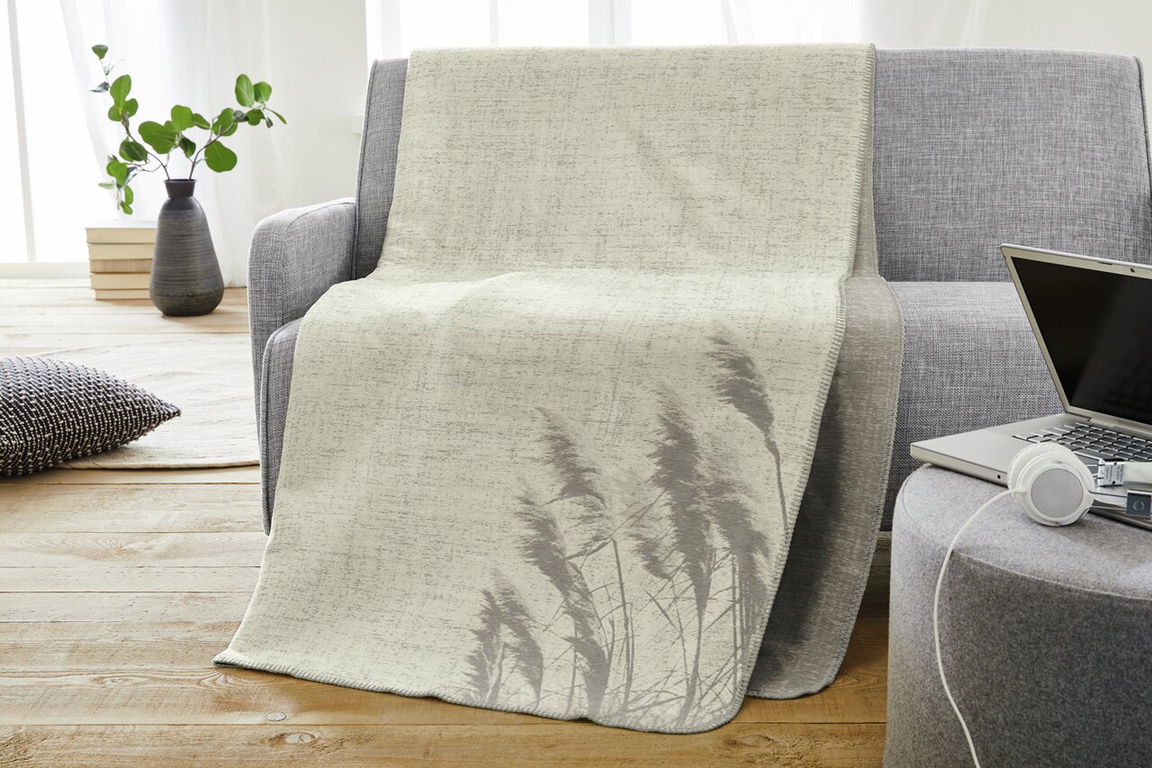 Jacquarddeken in lichtbeige met plantenmotief, gedrapeerd over een grijze bank in een moderne woonkamer, gezien vanuit de zijkant.