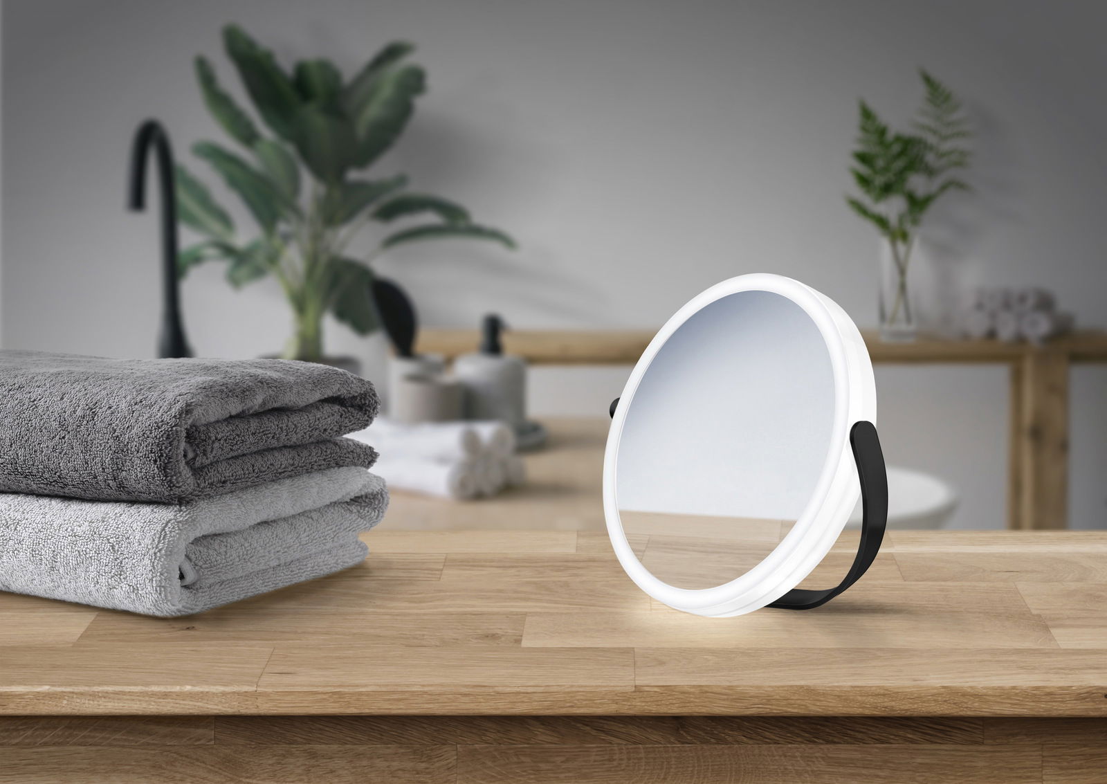 Schwarzer Kosmetikspiegel mit weißem Rahmen, auf einem Holzuntergrund stehend, im Hintergrund Handtücher und Pflanzen, seitliche Perspektive.