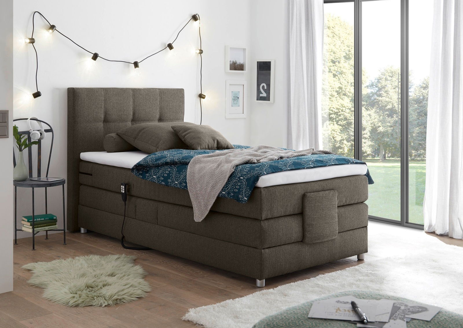 Boxspringbett in einem modernen Schlafzimmer, seitliche Perspektive mit dekorativen Kissen und Decke, umgeben von stilvollen Lichterketten und großen Fenstern.
