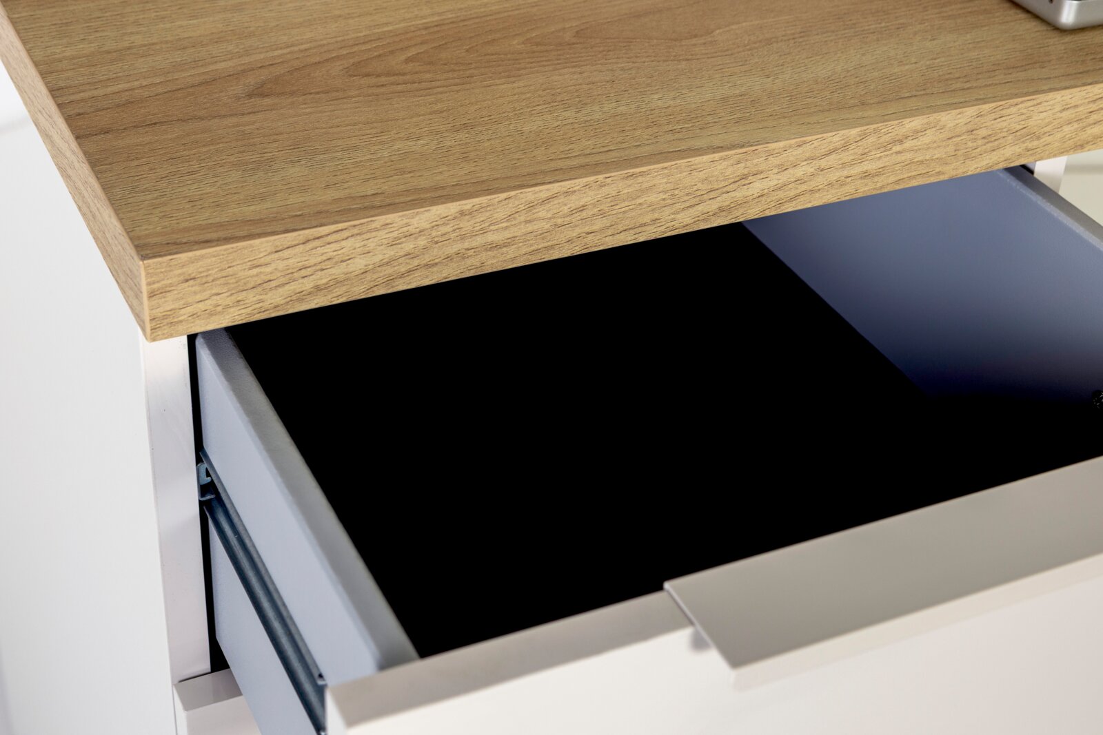 Nahaufnahme aus schräger Draufsicht: geöffnete Schublade eines modernen Möbelstücks mit weißer Front und Holzplatte in Eichenoptik