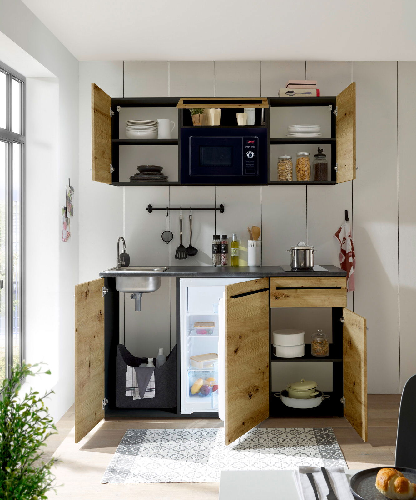 Frontale Ansicht einer kompakten Single-Küche mit Holzfronten, integriertem Kühlschrank, Spüle und Mikrowelle, ideal für kleine Räume.