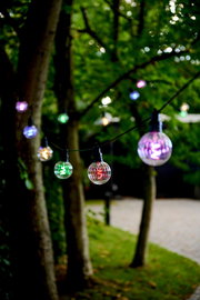 Bunte Lichterkette mit 10 LED-Lampen, aufgehängt zwischen Bäumen in einem Garten, aus der Nähe fotografiert.