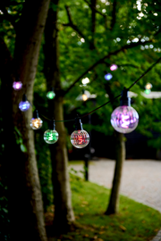 Bunte Lichterkette mit 10 LED-Lampen, aufgehängt zwischen Bäumen in einem Garten, aus der Nähe fotografiert.