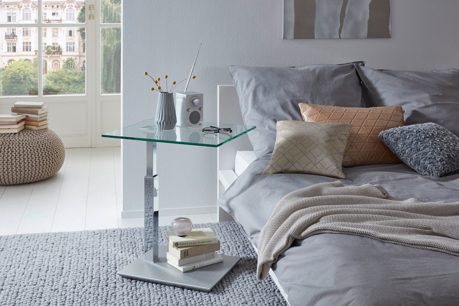 Moderner Couchtisch mit Glasplatte und Metallfuß, seitliche Perspektive, neben einem Bett mit grauer Bettwäsche und Kissen.