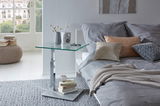 Moderner Couchtisch mit Glasplatte und Metallfuß, seitliche Perspektive, neben einem Bett mit grauer Bettwäsche und Kissen.
