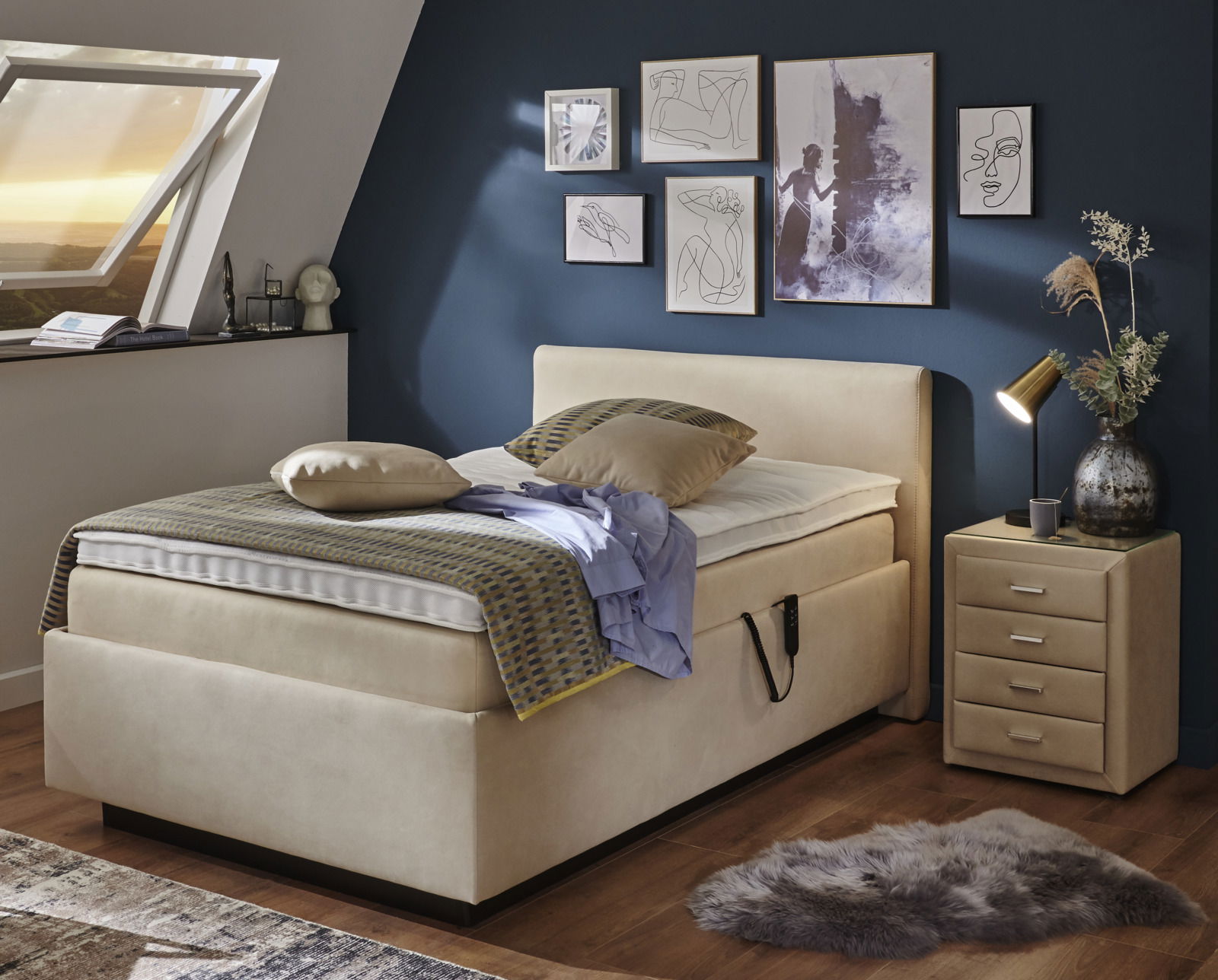 Gemütliches Polsterbett in einem stilvoll eingerichteten Schlafzimmer mit blauer Wand, dekorativen Bildern und einem Nachttisch. Perspektive von der Seite.