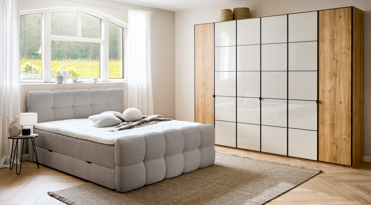 Modernes Boxbett in einem hellen Schlafzimmer mit großem Fenster, grauem Polsterkopfteil und passendem Schrank, seitliche Perspektive.