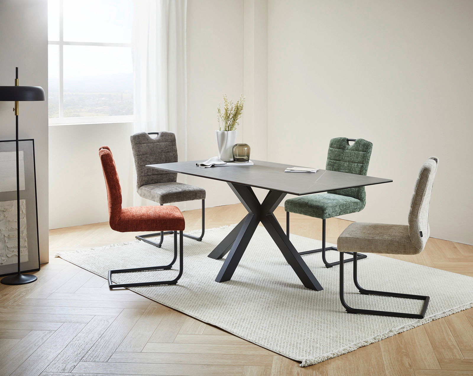 Moderner Esstisch mit schwarzem X-förmigem Gestell und grauer Tischplatte, umgeben von vier farbigen Stühlen in einem hellen Raum mit Fenster im Hintergrund, seitliche Perspektive.