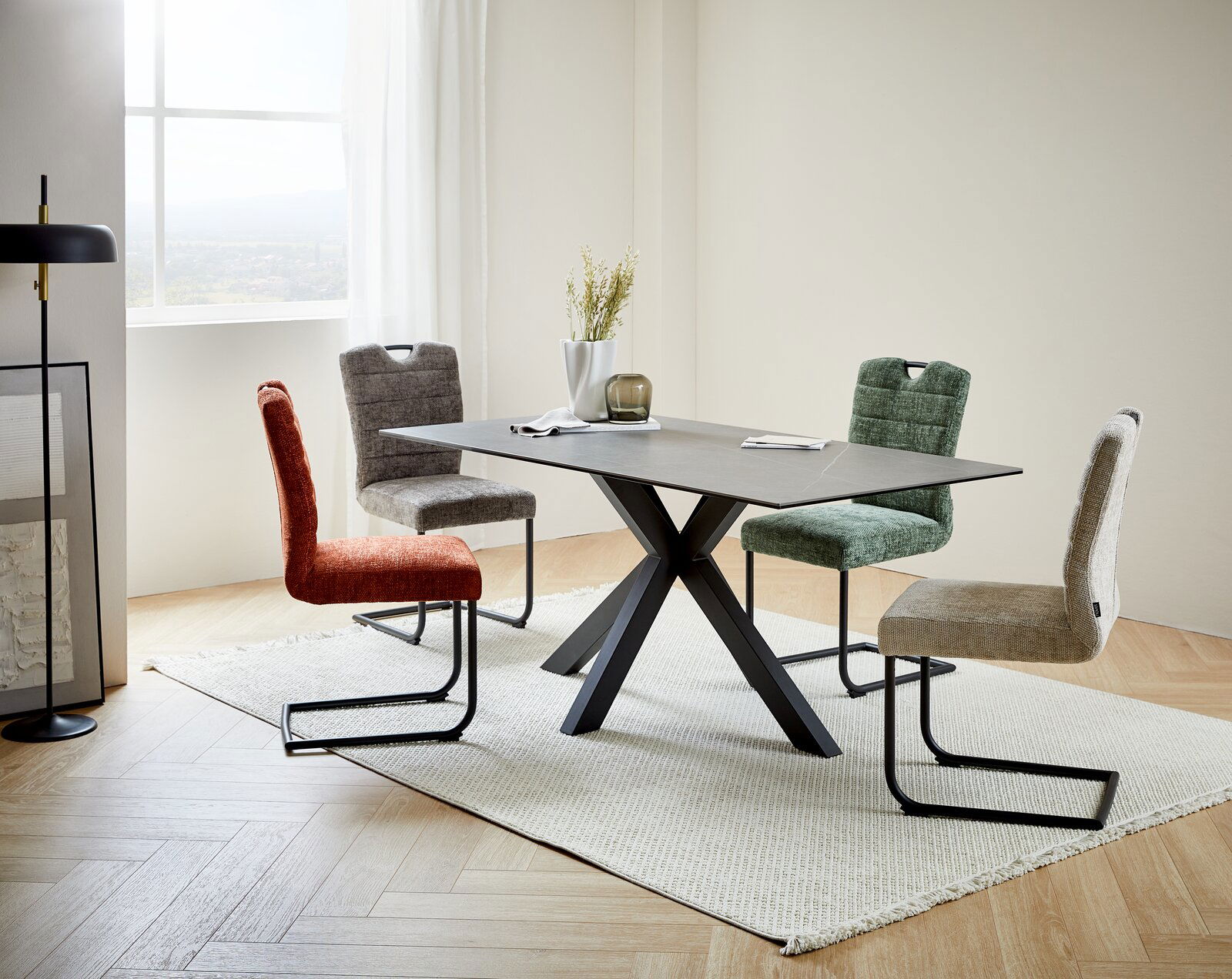Moderner Esstisch mit schwarzem X-förmigem Gestell und grauer Tischplatte, umgeben von vier farbigen Stühlen in einem hellen Raum mit Fenster im Hintergrund, seitliche Perspektive.