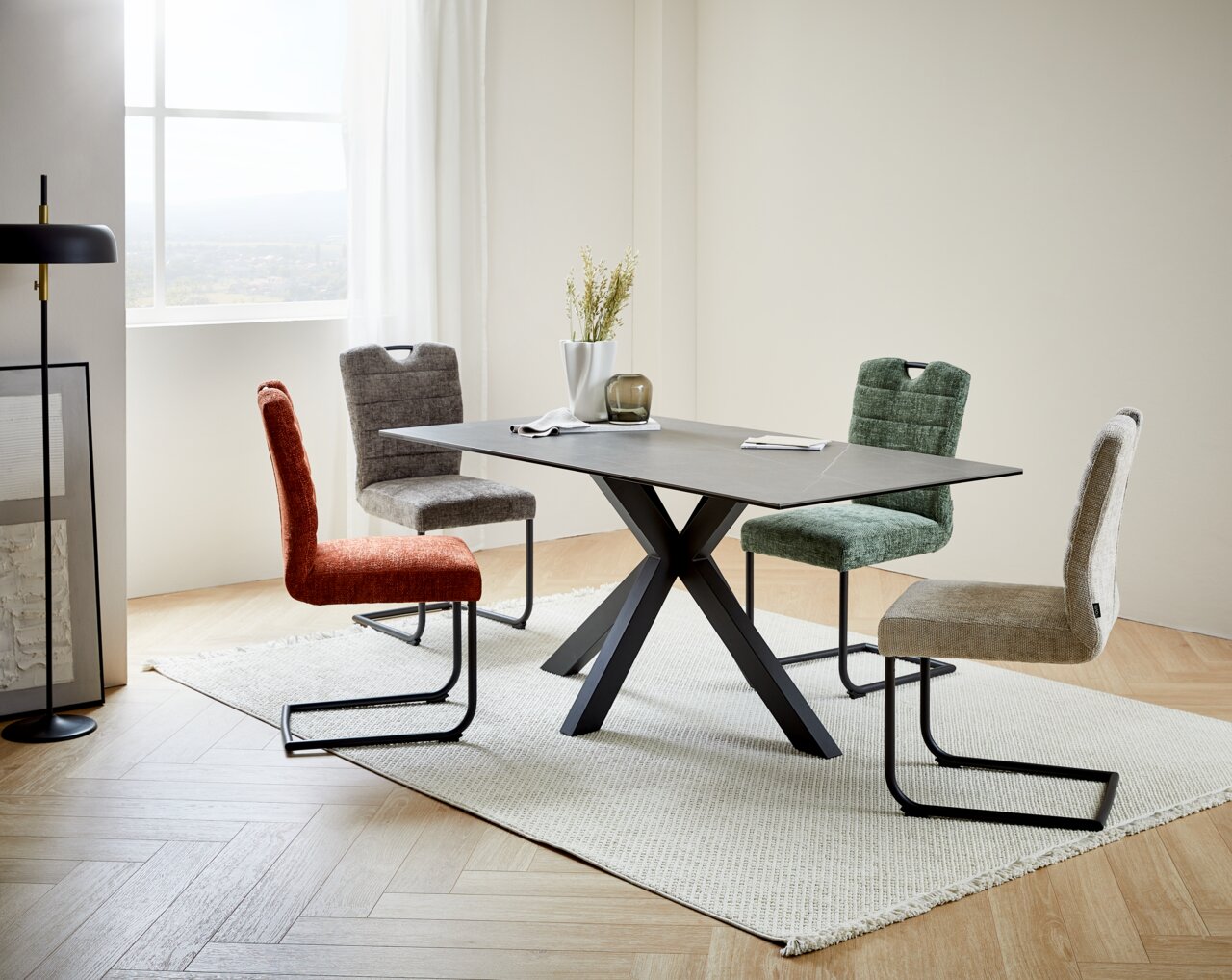 Moderne eettafel met een zwart X-vormig onderstel en een grijs tafelblad, omringd door vier gekleurde stoelen in een lichte ruimte met een raam op de achtergrond, gezien vanuit een zijdelingse hoek.