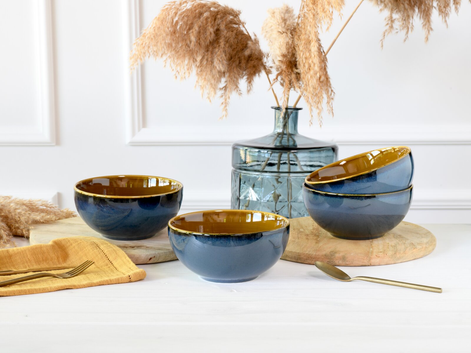 Creatable Buddha Bowl 4-tlg MALLORCA Drei glasierten Keramikschalen in Blau mit senfgelber Innenseite und Goldrand, eine Schale im Vordergrund und zwei rechts gestapelt, auf Holzbrett neben Glasvase mit Trockenblumen – Frontansicht in leichter Schrägperspektive.