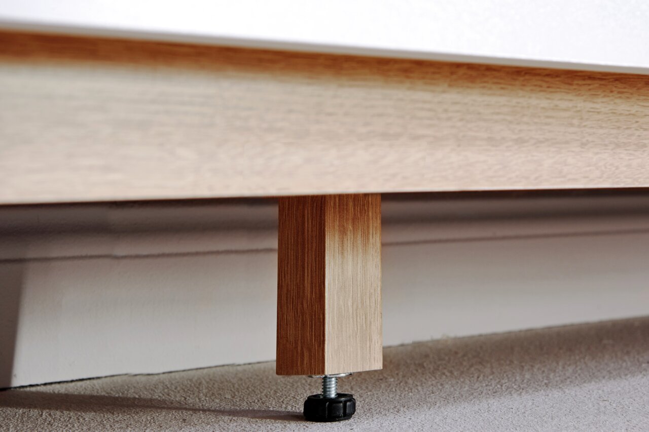 Nahaufnahme eines Holzfußes eines Sideboards mit verstellbarem Kunststofffuß auf Teppichboden.
