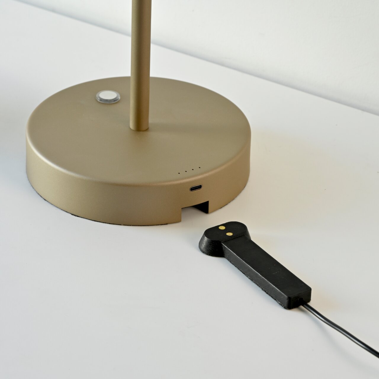 Nahaufnahme der Basis einer goldenen Akku-Tischleuchte mit USB-Anschluss und einem schwarzen Ladekabel auf einer weißen Oberfläche.