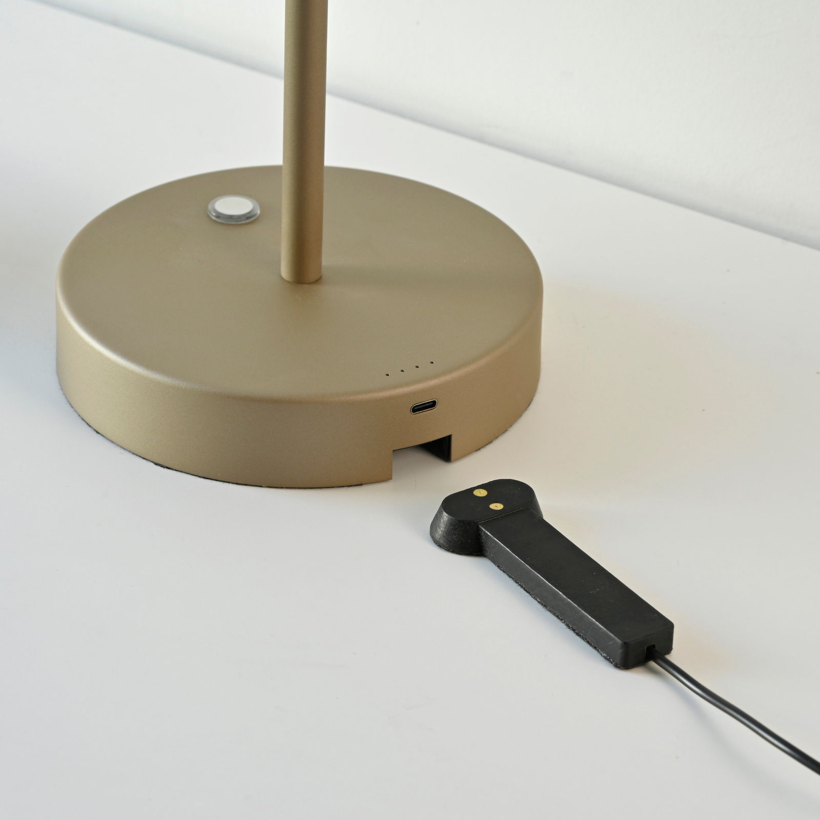 Nahaufnahme der Basis einer goldenen Akku-Tischleuchte mit USB-Anschluss und einem schwarzen Ladekabel auf einer weißen Oberfläche.