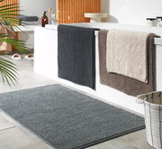 Badezimmer mit drei Badteppichen in verschiedenen Farben, die über den Rand einer Badewanne hängen, und einem grauen Badteppich auf dem Boden. Perspektive von schräg oben.