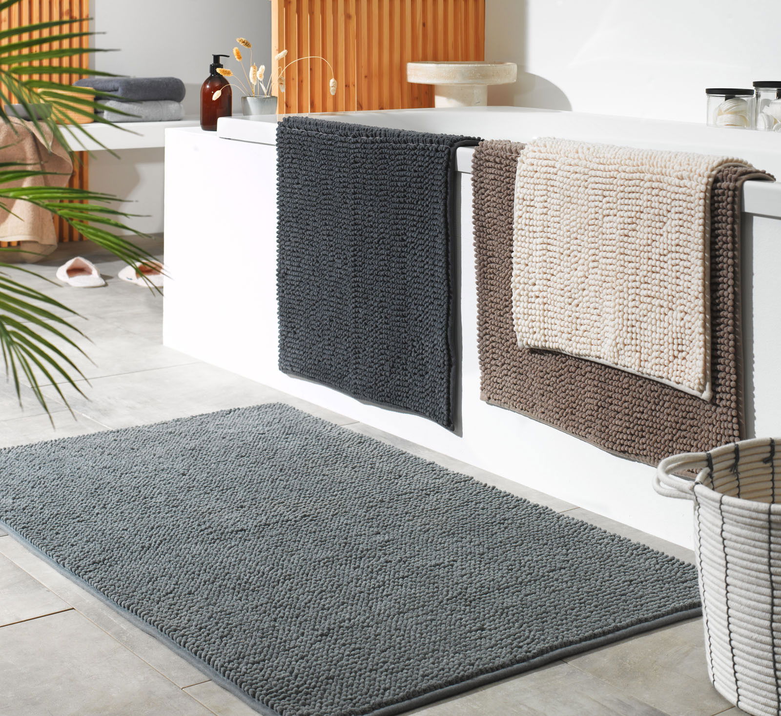 Badezimmer mit drei Badteppichen in verschiedenen Farben, die über den Rand einer Badewanne hängen, und einem grauen Badteppich auf dem Boden. Perspektive von schräg oben.