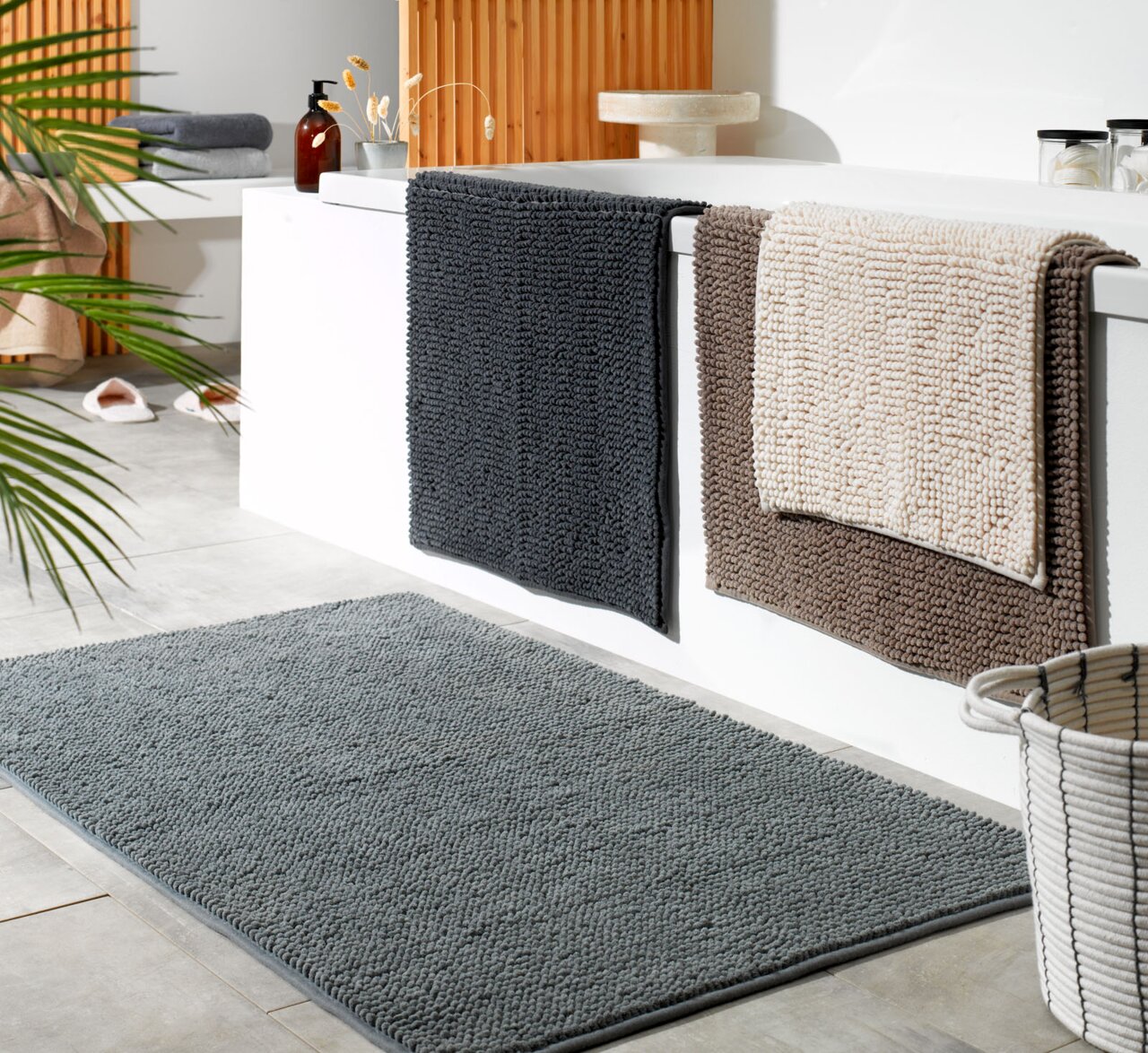 Badezimmer mit drei Badteppichen in verschiedenen Farben, die über den Rand einer Badewanne hängen, und einem grauen Badteppich auf dem Boden. Perspektive von schräg oben.