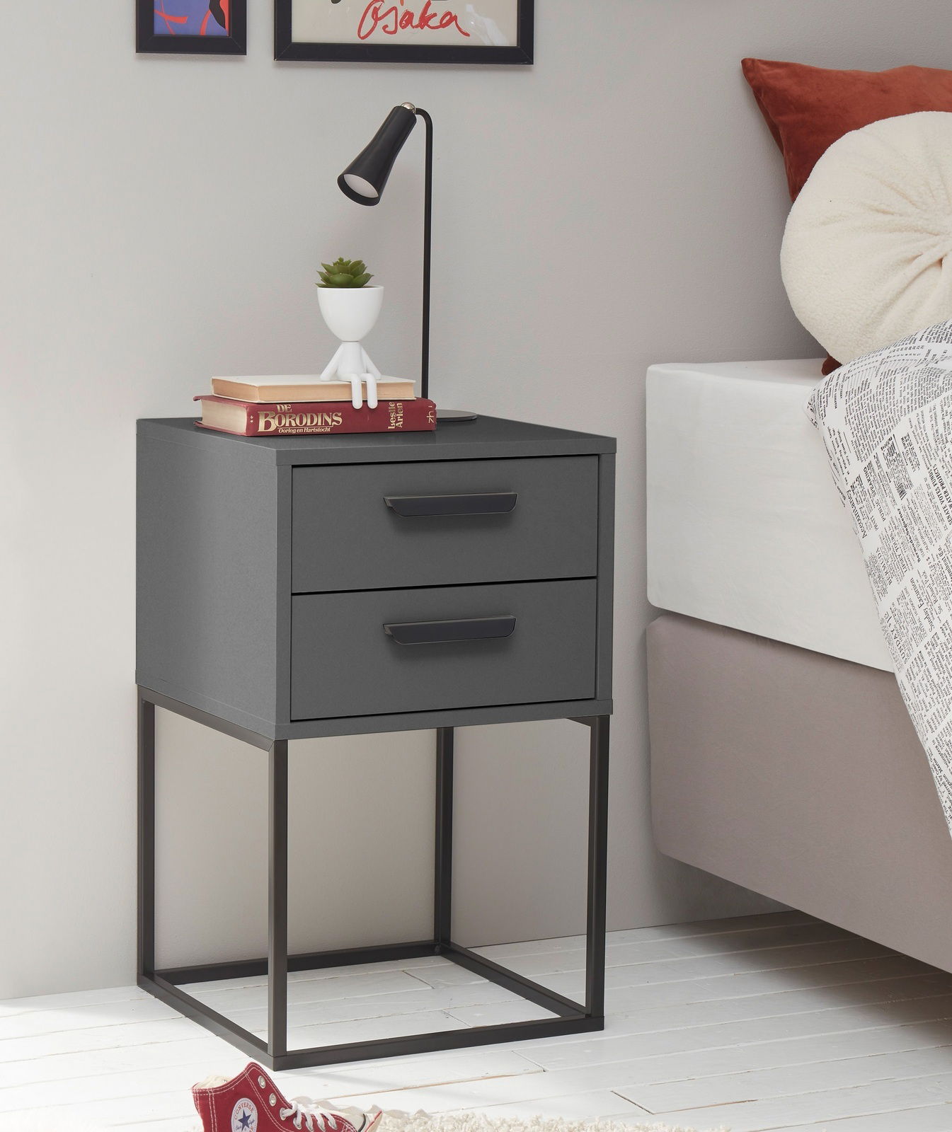 Moderne Nachtkommode mit zwei Schubladen in dunklem Grau, auf einem Metallgestell stehend, seitliche Perspektive. Dekoriert mit einer Lampe, Büchern und einer kleinen Pflanze.