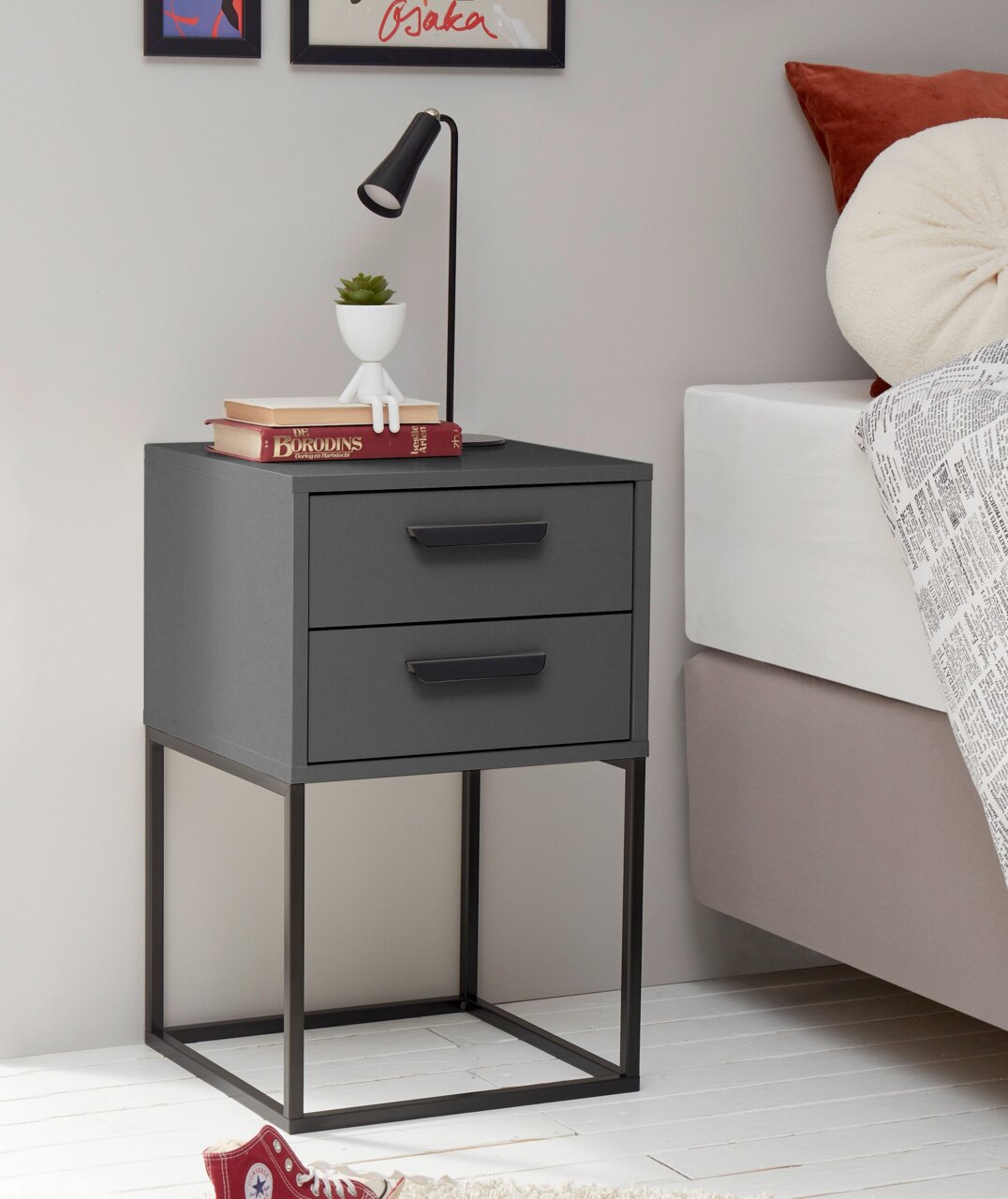 Moderne Nachtkommode mit zwei Schubladen in dunklem Grau, auf einem Metallgestell stehend, seitliche Perspektive. Dekoriert mit einer Lampe, Büchern und einer kleinen Pflanze.