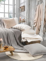 Gemütliche Wohnzimmerszene mit verschiedenen Fellkissen und Decken in neutralen Tönen, arrangiert auf einem Sofa und einem Korb, mit Kerzen und Dekoration im Hintergrund. Perspektive von vorne.