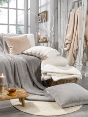 Gemütliche Wohnzimmerszene mit verschiedenen Fellkissen und Decken in neutralen Tönen, arrangiert auf einem Sofa und einem Korb, mit Kerzen und Dekoration im Hintergrund. Perspektive von vorne.