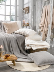 Gemütliche Wohnzimmerszene mit verschiedenen Fellkissen und Decken in neutralen Tönen, arrangiert auf einem Sofa und einem Korb, mit Kerzen und Dekoration im Hintergrund. Perspektive von vorne.