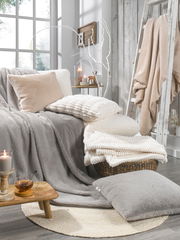 Gemütliche Wohnzimmerszene mit verschiedenen Fellkissen und Decken in neutralen Tönen, arrangiert auf einem Sofa und einem Korb, mit Kerzen und Dekoration im Hintergrund. Perspektive von vorne.