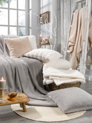 Gemütliche Wohnzimmerszene mit verschiedenen Fellkissen und Decken in neutralen Tönen, arrangiert auf einem Sofa und einem Korb, mit Kerzen und Dekoration im Hintergrund. Perspektive von vorne.