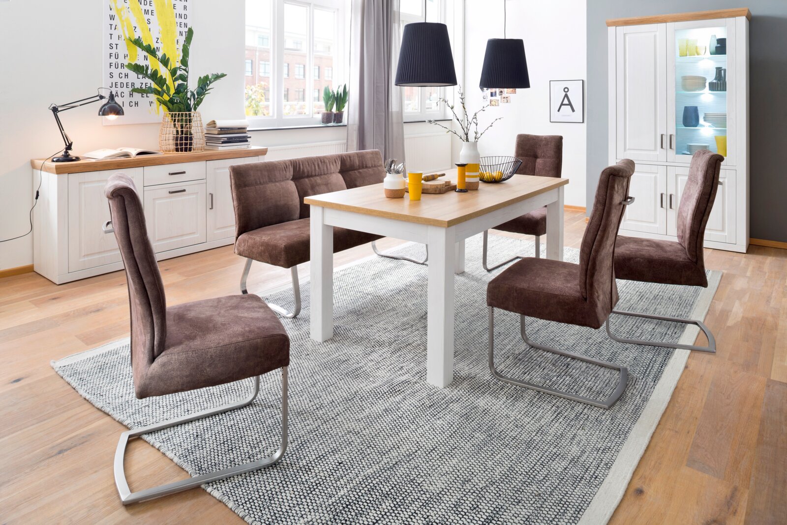 Modernes Esszimmer-Set mit weißem Holztisch und Holzoptik-Platte, brauner Polsterbank und vier braunen Schwingstühlen auf grauem Teppich, fotografiert in leicht erhöhter Frontalansicht.