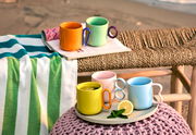 Bunte Kaffeebecher in verschiedenen Farben auf einem Tablett, umgeben von einem Handtuch und Zeitschriften, mit einem Strand im Hintergrund. Perspektive von vorne.