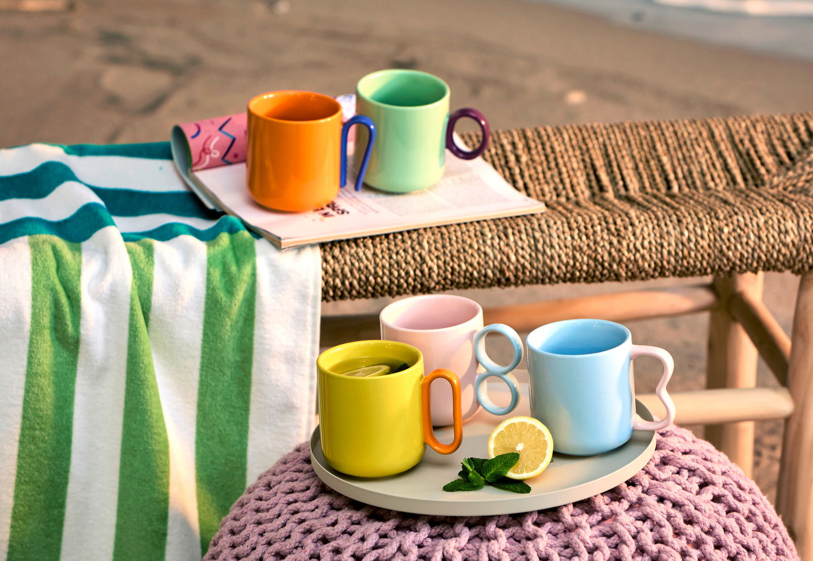 Bunte Kaffeebecher in verschiedenen Farben auf einem Tablett, umgeben von einem Handtuch und Zeitschriften, mit einem Strand im Hintergrund. Perspektive von vorne.