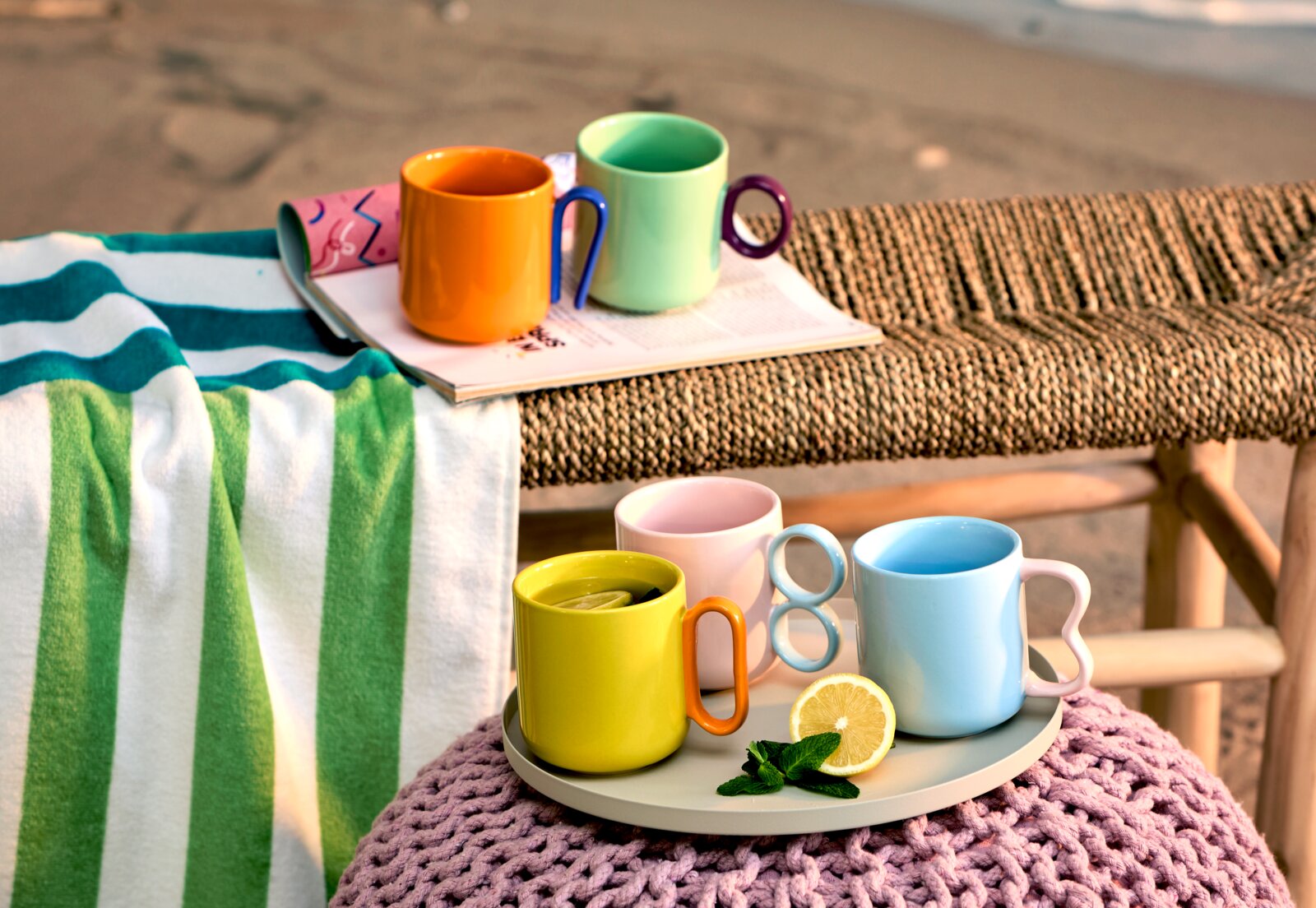 Kleurrijke koffiemokken in verschillende kleuren op een dienblad, omringd door een handdoek en tijdschriften, met een strand op de achtergrond. Vooraanzicht.