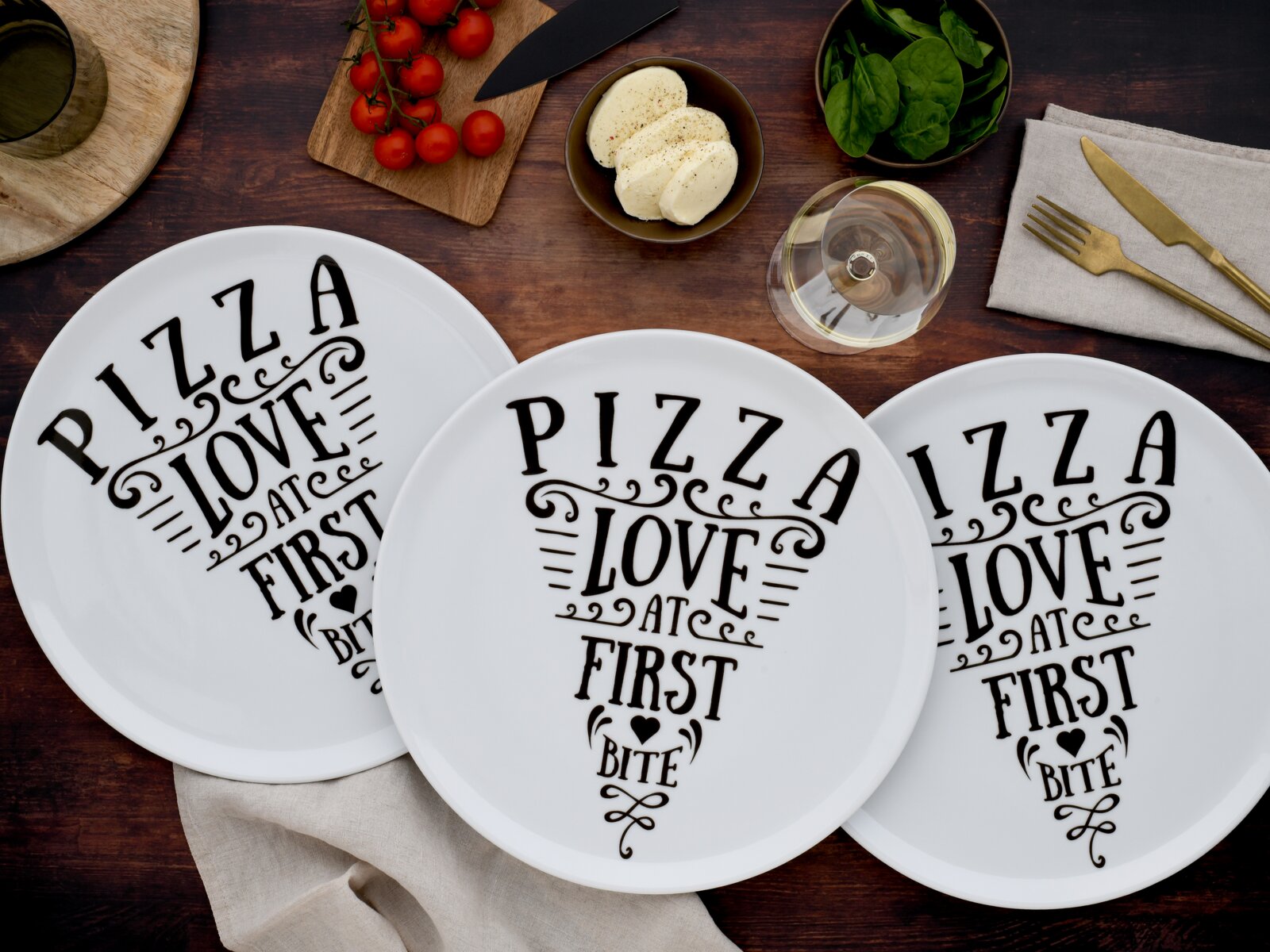 Drei weiße Pizzateller mit schwarzem Schriftzug „PIZZA LOVE AT FIRST BITE“ auf rustikalem Holztisch, Draufsicht von oben.