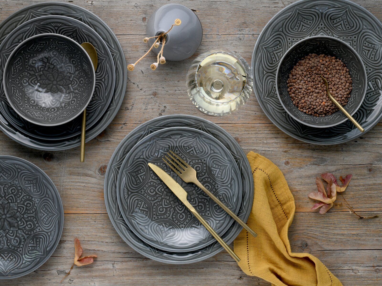 Dunkelgraues Geschirrset mit geprägtem Ornamentmuster (Teller und Schalen) auf Holztisch, mit goldfarbenem Besteck, Glas und gelbem Stofftuch, Draufsicht von oben