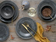 Dunkelgraues Geschirrset mit geprägtem Ornamentmuster (Teller und Schalen) auf Holztisch, mit goldfarbenem Besteck, Glas und gelbem Stofftuch, Draufsicht von oben