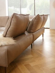 Braune Polsterrundecke mit großen Kissen, seitliche Perspektive, auf Parkettboden in einem hellen Raum.