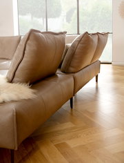 Braune Polsterrundecke mit großen Kissen, seitliche Perspektive, auf Parkettboden in einem hellen Raum.