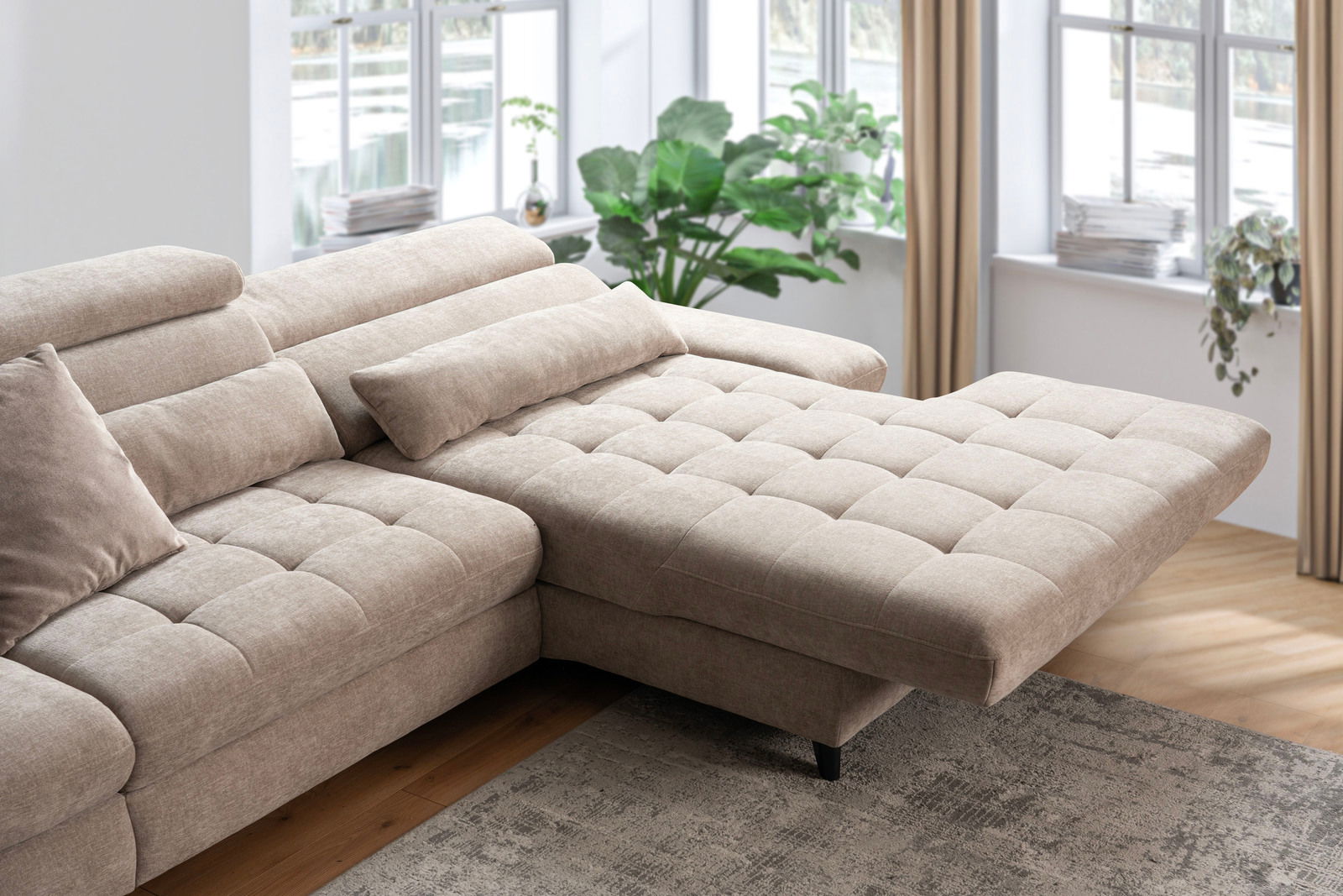 Beige Ecksofa mit verstellbaren Kopfstützen und einer Chaiselongue, fotografiert aus einer leicht erhöhten Perspektive in einem hellen Wohnzimmer mit großen Fenstern und Pflanzen.