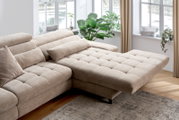 Beige hoekbank met verstelbare hoofdsteunen en een chaise longue, gefotografeerd vanuit een iets hoger standpunt in een lichte woonkamer met grote ramen en planten.