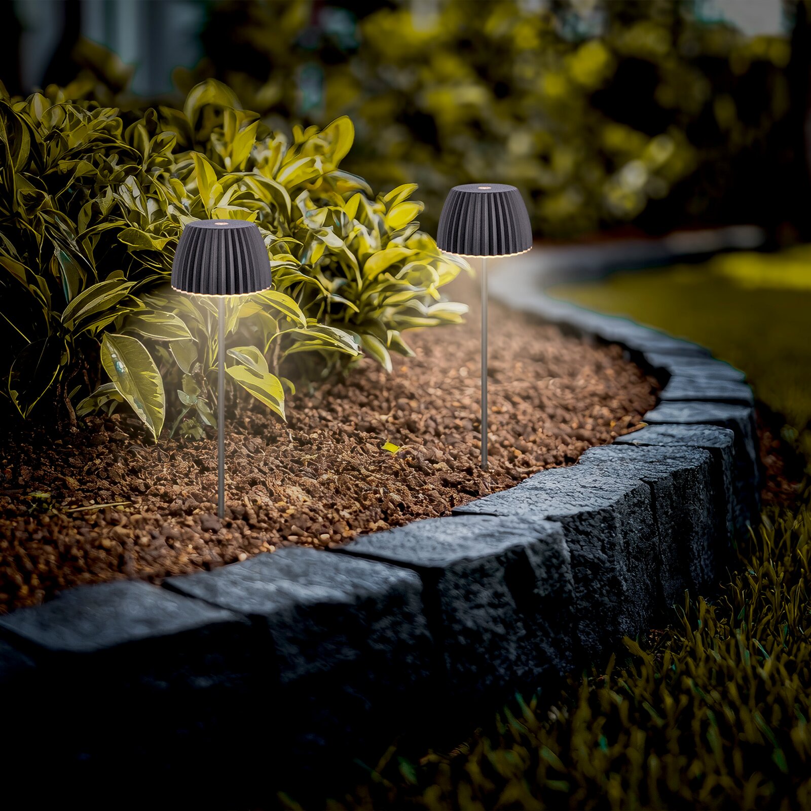 Zwei schwarze LED-Solar-Gartenleuchten mit geriffeltem Schirm auf Metallspieß, warmweiß leuchtend im Blumenbeet entlang einer Steinrabatte, aufgenommen aus niedriger Perspektive auf Augenhöhe der Leuchten