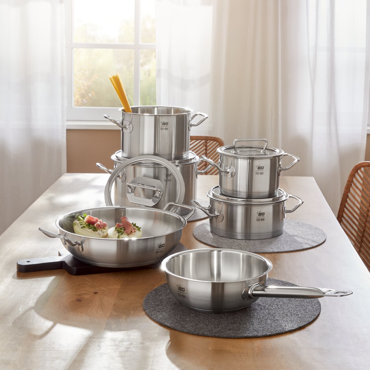 Topfset Gourmet Plus 6-tlg. aus Edelstahl, bestehend aus mehreren Töpfen und Pfannen, auf einem Tisch in einer hellen Küche präsentiert. Perspektive von vorne.