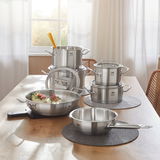 Topfset Gourmet Plus 6-tlg. aus Edelstahl, bestehend aus mehreren Töpfen und Pfannen, auf einem Tisch in einer hellen Küche präsentiert. Perspektive von vorne.