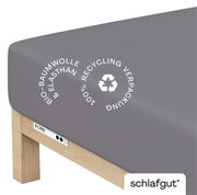 Grijs hoeslaken van biologisch katoen en elastaan, gefotografeerd vanaf een schuine hoek bovenop een matras, met vermelding van 100% gerecyclede verpakking.