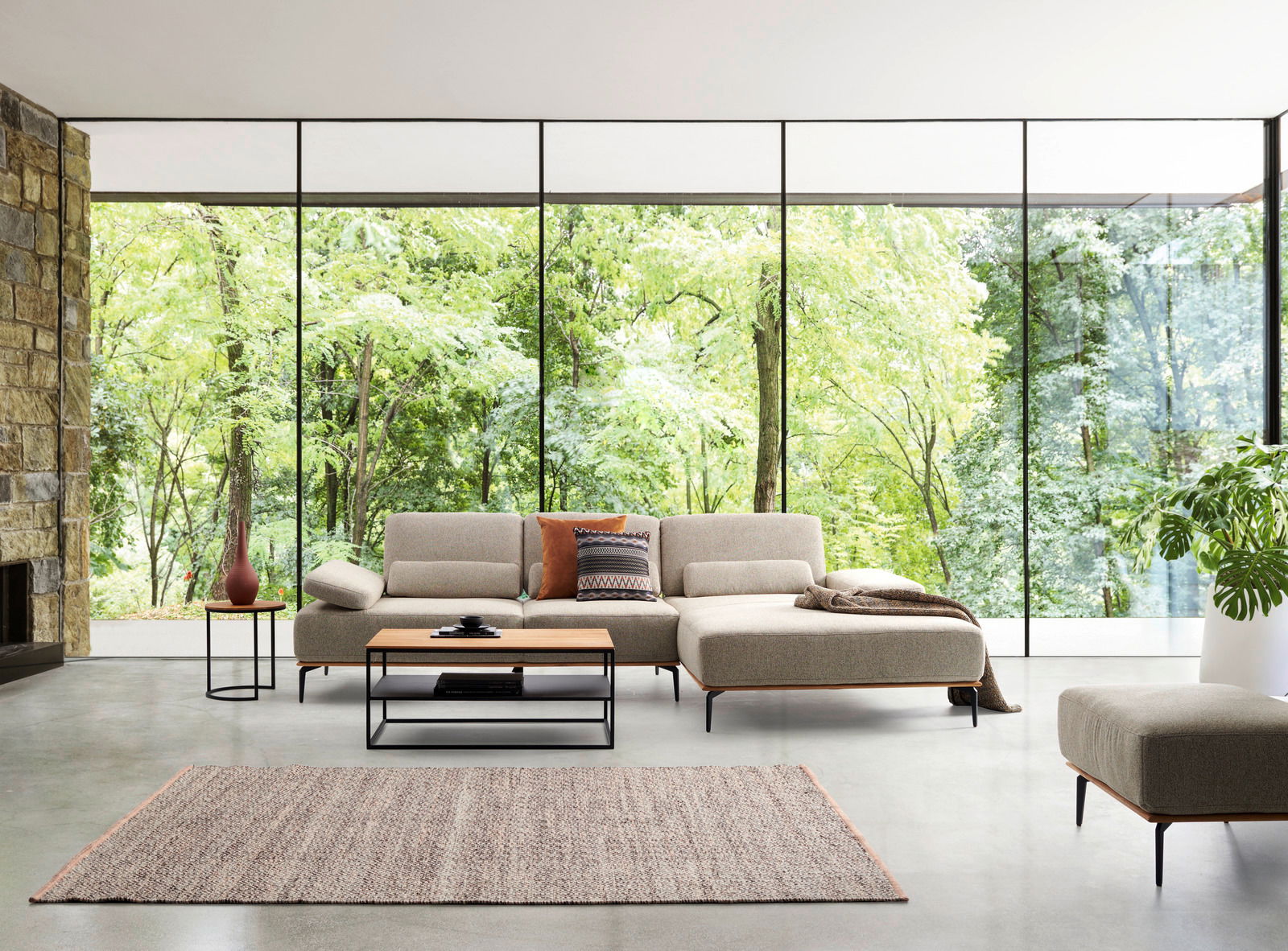 Moderne Longchairkombination in einem hellen Raum mit großen Fenstern, die einen Blick auf einen grünen Wald bieten. Die Perspektive zeigt die Frontansicht des Sofas mit einem Couchtisch und einem Beistelltisch.