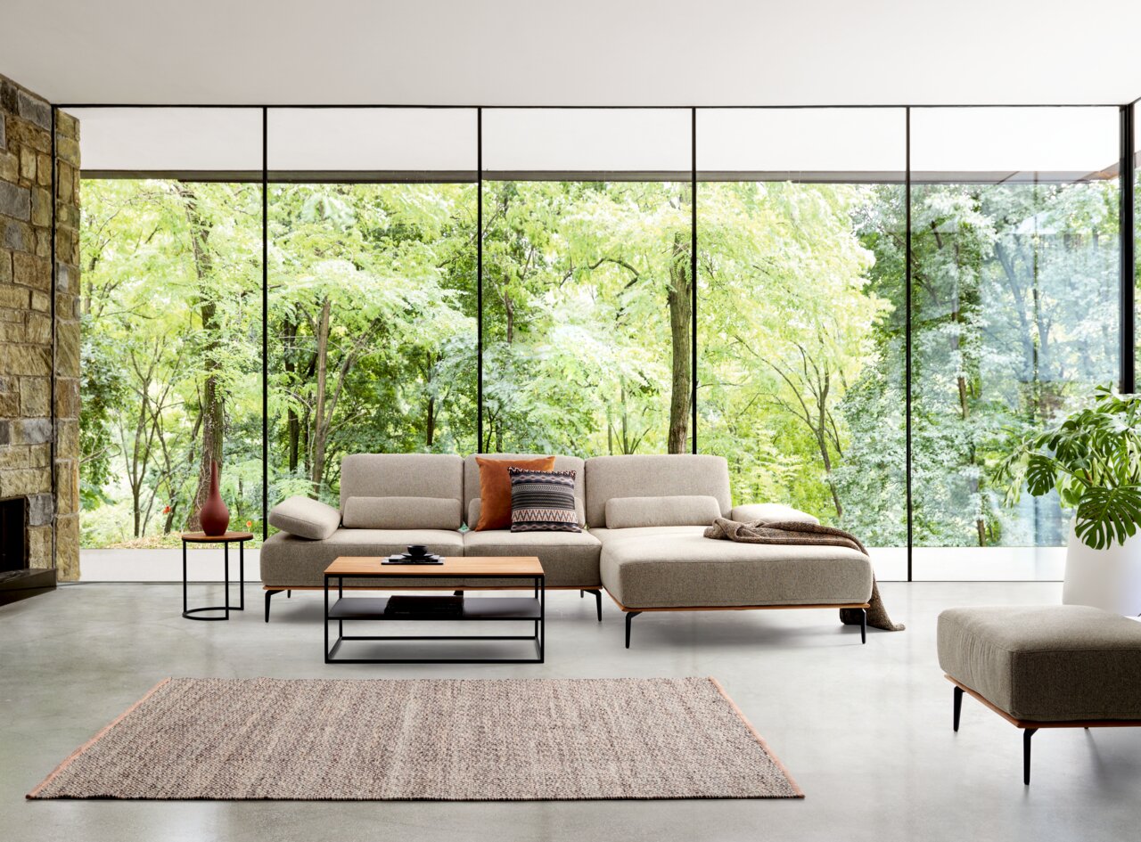Moderne Longchairkombination in einem hellen Raum mit großen Fenstern, die einen Blick auf einen grünen Wald bieten. Die Perspektive zeigt die Frontansicht des Sofas mit einem Couchtisch und einem Beistelltisch.