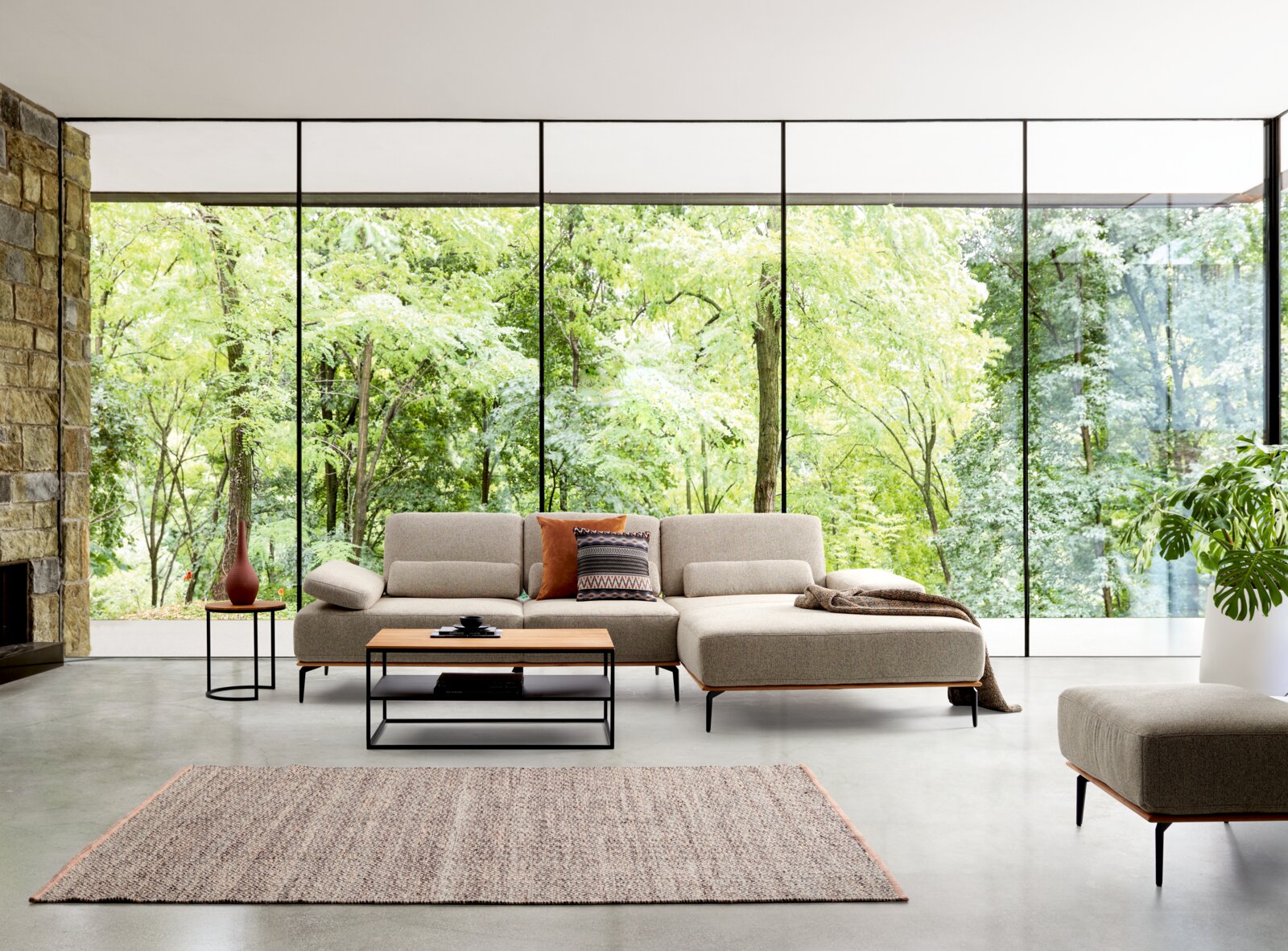 Valnatura Longchair-combinatie SPRINT Een moderne bankstelcombinatie in een lichte ruimte met grote ramen die uitzicht bieden op een groen bos. Het perspectief toont de voorkant van de bank met een salontafel en een bijzettafel.